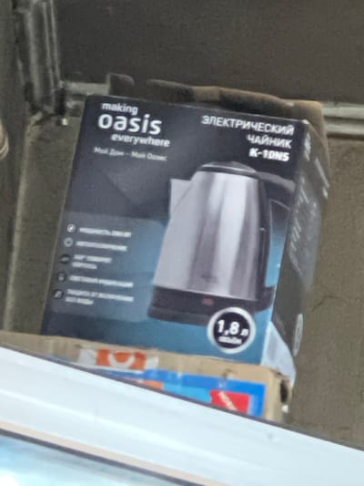 The image shows a close-up of a dark cardboard box that appears to be for an electric kettle. The box is tilted at an angle, making it difficult to see the entire object clearly.
The prominent text on the box is in Russian and English. In Russian, it reads "ЭЛЕКТРИЧЕСКИЙ ЧАЙНИК K-10NS," which translates to "ELECTRIC KETTLE K-10NS." Below this, there's text that appears to be a slogan or brand name, "making oasis everywhere," and further down, "Мой Дом - Мой Озис," meaning "My Home - My Oasis."
On the side of the box, there is a circular graphic with the number "1,8" inside, likely indicating the volume of the kettle in liters. A portion of another box or packaging is visible to the right, with the word "Нови" (Novi), possibly meaning "New" or part of a brand name.
The background is an out-of-focus, textured surface, possibly a wall or rough concrete. The lighting suggests it might be daytime, with some natural light illuminating the scene. The scene appears to be set in an outdoor or semi-outdoor location, given the rough textures and the way the box is placed. There are no people visible in the image. The overall impression is that of product packaging left or stored in a somewhat utilitarian setting.