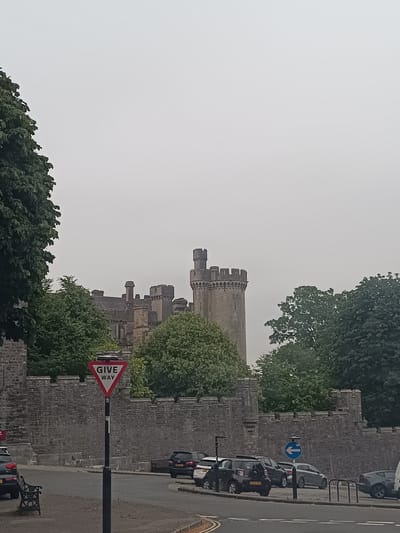 The image shows a view of Arundel Castle, a medieval castle in Arundel, West Sussex, England. The castle is a Grade I listed building and is one of the largest inhabited castles in the UK. In the foreground, there is a "Give Way" sign on a pole, and several cars are parked on the side of the road. There is also a bench visible. The sky is overcast, and there are trees and foliage surrounding the castle. The castle itself is a large stone structure with turrets and battlements, and it appears to be in good condition.