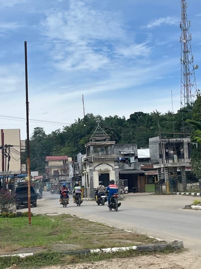 The image captures a street scene in Alurcucur, Indonesia, under a partly cloudy sky. In the foreground, a rough, sandy area with sparse grass and a low concrete barrier with black and white stripes borders a paved road. The road itself has a gray asphalt surface with white lane markings visible.
Several people on motorcycles are moving away from the viewer. The closest motorcyclist on the right wears a light-colored shirt and a dark helmet. To their left, a motorcyclist in a blue and red jacket with a white stripe and a dark helmet is riding. Further ahead, another motorcyclist wearing a green and white striped shirt and a dark helmet is visible, followed by a motorcyclist in a dark helmet and a yellow vest. The motorcycles appear to be of a common scooter or underbone style.
On the left side of the road, a black pickup truck is parked. Adjacent to it, a tall, dark pole stands, possibly a utility pole. Behind the truck, a building with a light-colored facade and some signage is visible. Further down the street, on the right side of the road, a multi-story building with a distinctive architectural style, featuring a decorative upper section with pointed elements, is prominent. Behind these buildings, dense green foliage and trees cover a sloping hillside.
Dominating the right side of the background is a tall, red and white lattice telecommunications tower. The overall impression is a moderately populated urban or semi-urban environment in Indonesia, with ongoing activity on the street.