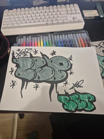 The image displays a close-up, overhead view of a white sheet of paper with graffiti art on it, positioned on a dark surface that appears to be a desk. To the top of the paper, a white keyboard is visible. In the background, to the right of the keyboard, a pack of HEETS and an IQOS device are present. The graffiti on the paper consists of two distinct pieces. The larger piece, in the center, is outlined in black and filled with a light green color, featuring bubble-like letters with a few small star-like embellishments scattered around it. Above this larger piece, there are some black scribbled lines and symbols. Below the larger piece, to the right, is a smaller graffiti tag, also outlined in black and filled with green, appearing to be the letters "SRB". The drawing is situated on a flat surface, possibly a desk or table, with hints of a wooden floor visible at the bottom. The environment suggests an indoor setting, likely a workspace. There is no indication of the city of Novi han, Bulgaria.