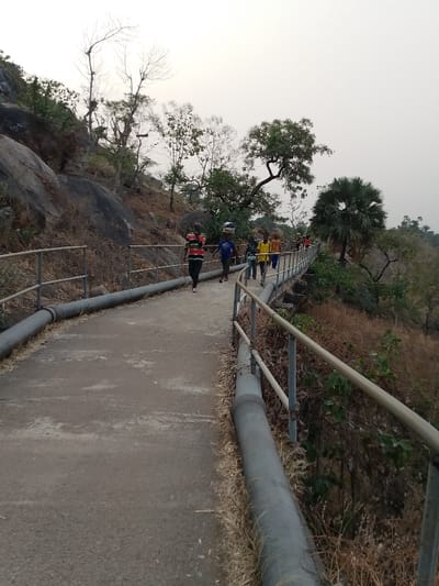 A group of people walks along a concrete path with metal railings and large pipes running alongside it. The path is built on a hillside with rocky terrain and dry vegetation. Some trees with bare branches are visible along the path, as well as a tall palm tree. The sky is overcast and creates a muted light.