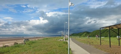The image depicts a coastal scene in Aleksandrovsk-Sakhalinskiy, Russia. In the foreground, a paved walkway runs alongside a grassy area. Beyond the walkway, a sandy beach stretches towards the ocean, with a calm sea meeting the shore. In the distance, a line of green hills forms the backdrop, partially obscured by dramatic, dark storm clouds gathering in the sky. A few scattered small buildings and vehicles are visible on the grassy area near the hills. A single lamppost with security cameras is prominently positioned on the walkway. Several people can be seen walking on the beach in the distance, and a vehicle is parked on the sand near the edge of the grass. A covered seating area with a tan canopy is situated in the mid-ground to the right.