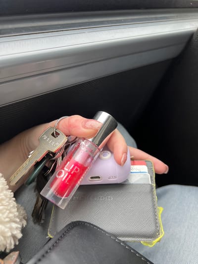 This image depicts a close-up view of a person's hand inside a vehicle in the city of Липицы, Russia. The hand, exhibiting light-colored, possibly glittery, nail polish, is holding a keyring with several keys, including a prominent brass-colored key labeled "APEX." Directly below the fingers, a clear cylindrical bottle containing red liquid, labeled "essence LIP OIL," is held upright. The hand and the held objects rest upon a dark gray textured wallet or cardholder, from which a bright yellow string or tag is partially visible, along with multiple cards inserted into its slots. Partially obscured behind the lip oil bottle and resting on the wallet is a small, light purple electronic device with a paw print design on its top surface and a USB-C port at its base. The person is wearing light blue jeans, visible in the lower right of the frame, and a fluffy white material is present in the lower left. The background shows the interior of a vehicle, specifically a car door panel with a light grey trim and a dark textured surface.