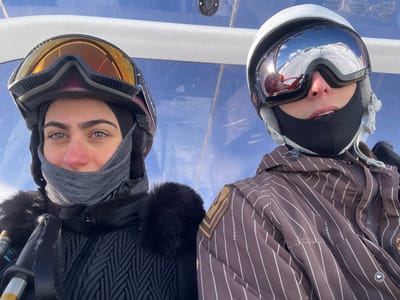Two individuals are depicted in a close-up, eye-level shot, likely within a ski lift cabin, against a clear blue sky background. The scene is located in Valtournenche, Italy.
The individual on the left, identified as female, has a fair complexion, dark hair, dark eyebrows, and light-colored eyes. She wears a dark ski helmet with amber-tinted ski goggles pushed onto the helmet, revealing her upper face. A grey neck gaiter covers her lower face, leaving her eyes visible. She is dressed in a black quilted ski jacket with a prominent black faux-fur collar. A portion of a ski pole is visible near her left shoulder.
The individual on the right wears a white ski helmet and large, reflective ski goggles that obscure their face. The goggles reflect a bright sun and a snow-covered mountain landscape. A black face mask or balaclava covers their lower face. This individual is wearing a brown ski jacket with thin white vertical stripes and dark shoulder patches. The upper portion of a ski pole is visible near their right shoulder.
Both individuals are dressed in winter ski attire, indicating an outdoor, cold weather environment. The lighting suggests a bright, sunny day.