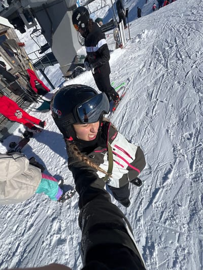 The image displays a sunny winter day on a snow-covered ski slope in Tignes, France. The primary subject, a female, is positioned in the foreground, taking a low-angle selfie. She wears a black ski helmet with Uvex goggles, a white and pink Rehall brand ski jacket, black ski pants, and black ski boots. Her long blonde hair is braided and visible below the helmet. Her expression appears to be a grimace.
Behind and to the left of the main subject, another individual stands on skis, wearing a black POC helmet, a black ski jacket with white horizontal stripes, and black ski pants. This person is looking downwards, possibly adjusting equipment. Further to the left, a grey metallic ski lift structure is partially visible, alongside a wooden building. A person in red ski attire is partially visible near the building, and a red jacket is draped over an unseen object. In the bottom-left corner, the lower body of a third person is visible, wearing light beige pants and multi-colored (mint green, pink, purple) ski pants, with black ski boots on a snowboard or skis. The snow-covered terrain features numerous ski tracks. In the upper background, additional skiers and distant mountains are visible under a clear, bright sky.