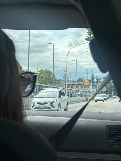 The image shows a view from inside a car, looking out at a street scene in Ascoli Piceno, Italy. In the foreground, the back of a person's head and shoulders are visible, wearing sunglasses. The person appears to be looking towards the right. Through the windshield, several cars are driving on a road. One white car in the middle ground has its headlights on and a license plate that appears to be "ES 349RY". In the background, there are buildings, trees, and streetlights. A yellow sign with the Lidl logo is visible on a building in the distance. The sky is cloudy. The overall impression is a typical urban street scene on an overcast day.