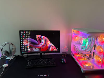 A well-lit, frontal shot depicts a computer desk setup. The setup includes a monitor displaying a colorful abstract image, a black keyboard, a black gaming mouse, a white and grey headset on a stand, and white earbuds. To the right of the monitor is a white computer tower with a transparent case, illuminated by multicolored LED lights. A black Huzaro mousepad covers most of the desk's surface. The wall behind the desk is a neutral color, suggesting an indoor environment. There is no indication of the image's location being in Dragor, Bulgaria.