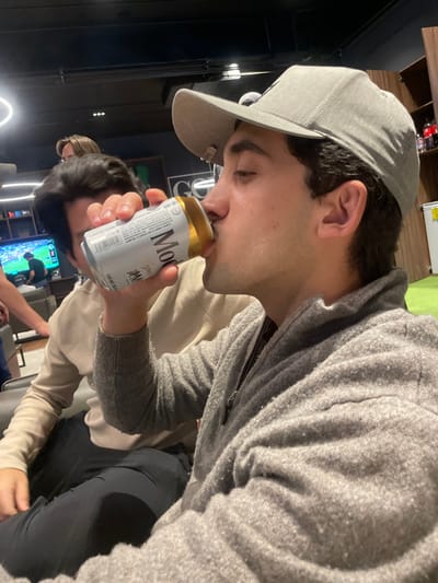 An indoor scene is depicted in La Purísima, Mexico, featuring a group of individuals in a casual setting. In the foreground, a male subject, wearing a grey baseball cap backward and a grey zippered fleece jacket, is observed drinking from a silver and gold "Modelo Especial" beer can. He holds the can to his mouth with his right hand, his profile visible. Behind him, a second male is seated, wearing a light-colored long-sleeved shirt, with his face partially obscured by the foreground subject and the can. Additional individuals are partially visible in the background: a third person's head behind the second male, and an arm and leg belonging to a fourth person on the far left. The environment consists of dark walls and ceiling with modern lighting fixtures. A flat-screen television on the left displays what appears to be a soccer match. On the right, shelving holds various bottles, and a white appliance, possibly a refrigerator, is visible. A large "G" logo is present on a dark wall section in the mid-right, and a section of light green flooring or rug is visible on the far right.