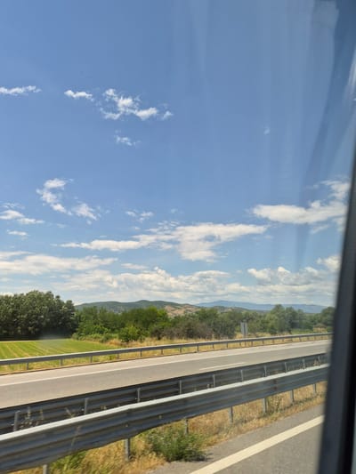The image depicts a view from a vehicle traveling on a highway, possibly in or near Chuchuligovo, Bulgaria. The scene is dominated by a bright blue sky with scattered white clouds. Below the sky, a landscape unfolds, featuring a highway with guardrails in the foreground and middle ground. The highway is paved and appears to be multi-lane. Beyond the highway, a green field is visible, bordered by a line of trees and shrubs. In the distance, rolling hills covered in vegetation and trees form the horizon, with a faint outline of mountains under the sky. The overall impression is a sunny day in a rural or semi-rural setting.