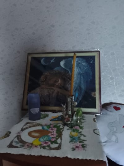 The media file shows a still life composition on a tabletop, likely indoors. The central focus is a framed picture, possibly a diamond painting, depicting an angelic figure with large blue wings against a dark, starry background with a crescent moon. To the left of the painting stands a tall, slender candle in a small metal holder that also contains a thermometer. In front of the painting, a purple cylindrical object, likely an air freshener, is placed on a white tablecloth with a colorful floral and egg design. Small decorative figurines, including a rabbit and a green abstract shape, are also visible on the table. To the right, a white pillow with a red heart and a stylized animal design peeks into the frame. The overall setting appears to be a domestic environment, with the patterned wallpaper in the background suggesting a casual interior. The lighting is soft, and there are no clear indicators of the time of day or weather conditions. The visible text on the purple object reads "AIR MAGIC FRESH AIR BLACK OCEAN." The composition evokes a sense of calm and decorative arrangement.