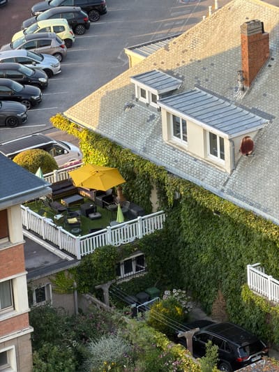 The image is an aerial view of a residential area in Dieppe, France, featuring a building with a prominent red brick chimney. The building has a grey tiled roof with several dormer windows. A section of the building is covered in dense green ivy. In front of this section, there is a white-railed patio area furnished with outdoor seating, including sofas, chairs, and a coffee table. A bright yellow umbrella is open on the patio. Adjacent to the building, a parking lot is visible with several cars parked in a row, including a silver hatchback, a light yellow car, and a black SUV. Further down, another building with brick and stone facade is partially visible, with greenery and flowering plants surrounding its base. A black SUV is parked in a lower area, partially obscured by vegetation. The lighting suggests it is late afternoon or early evening.