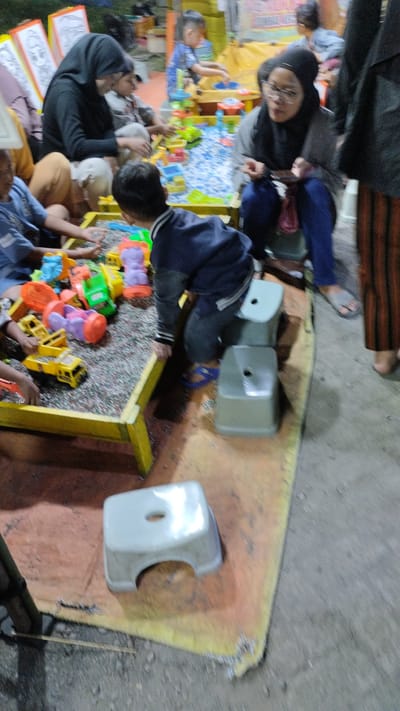 The image depicts a group of people, primarily children and a few adults, engaged in an activity at what appears to be an outdoor market or fair in Sukodono, Indonesia. The central focus is a sand table filled with numerous colorful plastic toys, including trucks, construction vehicles, and animal figures. Several children are actively playing with these toys.
In the foreground, a young boy wearing a dark blue jacket and gray pants is engrossed in the sand table. To his left, another child in a blue shirt is also playing with the toys. Behind the boy in the blue jacket, a woman wearing a black hijab and glasses is seated, looking down, possibly at her phone. Another woman in a black hijab is seated to the left of the sand table, also observing the children. In the background, a young child in a blue and green striped shirt is interacting with a colorful toy structure.
The scene is set on a textured yellow mat, with a dusty ground visible around it. Two small, light gray plastic stools are placed near the sand table. The lighting suggests it might be late afternoon or evening, as the light is somewhat dim and the scene is outdoors. There is no visible text in the image. The overall atmosphere seems to be one of casual recreation and community gathering.