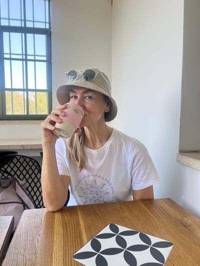 An adult female is seated at a light brown wooden table indoors, likely a cafe or restaurant in Rosh Haayin, Israel. She is wearing a beige bucket hat with round, clear-framed sunglasses perched on its brim, and a white t-shirt featuring a light blue graphic of a sun and text. Her light brown hair is visible beneath the hat. She is seen taking a drink from a light beige reusable cup with a pink silicone sleeve and lid, holding it with her right hand. Her gaze is directed towards the viewer with a slight smile. To her left, a large window with a dark grid frame overlooks green foliage and a bright sky. A portion of a dark, grid-patterned chair is visible to her left. A square coaster with a black and white geometric pattern rests on the table to her right. The background consists of a plain white wall.