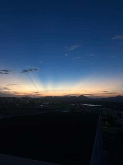 A vertical, eye-level shot captures a sunset scene. The left side of the frame shows a dark blue sky with some wispy clouds. As the view transitions towards the middle, the sky turns golden and orange, with visible sun rays shining downwards. On the right, a mountain range silhouettes against the bright sunset. In front of the mountains lies a city with buildings and streetlights. A large, dark structure is situated on the bottom right corner, obscuring part of the city view. The photo was taken in the city of Zibatá, Mexico.