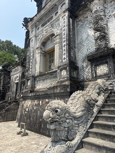 The image depicts a corner of a historic building, possibly a temple or palace complex, likely located in the city of Thủy Xuân Ward, Vietnam, judging by the architectural style and cultural details. The structure is characterized by intricate stone carvings, particularly a dragon-like figure that serves as a decorative element along a staircase. The building exhibits a blend of textures and patterns, with a prominent maze-like design on the ceiling and detailed inscriptions in a traditional script on the walls. A window with a lattice design can be observed along the façade. The overall ambiance suggests a place of historical and cultural significance, with the stone surfaces showing signs of age and weathering. Vegetation can be seen in the lower part of the image, hinting at an outdoor environment.