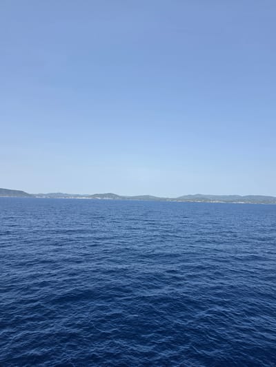 The image is a wide-angle shot of a coastline under a clear blue sky. The scene is divided horizontally, with the left side dominated by a vast expanse of light blue sky, suggesting a bright, sunny day. The right side features a deep blue sea with visible ripples and waves, indicating a slight choppiness on the water's surface.
Along the horizon, separating the sky and sea, is a long, low-lying coastline. This landmass appears mountainous and covered in vegetation, with a scattering of white buildings visible along the shore, suggesting a town or village. The buildings are small and blend into the landscape, with no prominent structures or landmarks immediately identifiable.
The time of day appears to be daytime, likely mid-morning or afternoon, given the brightness of the sky and the angle of the sun, which is not directly visible but implied by the illumination. The weather is clear and fair.
The image captures a serene and picturesque coastal landscape. The presence of the settlement along the coast suggests a populated area, likely a tourist destination given the typical features of such a scene. The image appears to be taken from a slightly elevated position, possibly a boat or a higher vantage point on the shore, looking out towards the sea and the land. There is no text visible in the image. The overall mood evoked is one of peace and natural beauty.