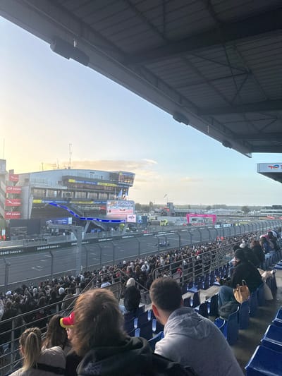 The image captures an active motor racing event at the Circuit de la Sarthe in Le Mans, France, viewed from an elevated grandstand during late afternoon or early evening. The scene is illuminated by a low sun, casting a soft, warm light on the left side of the frame.
In the immediate foreground, the underside of a dark metal grandstand roof structure, supported by exposed beams, dominates the upper portion of the image. Cylindrical speakers are mounted on these beams. Below the roof, blue stadium seats are heavily populated by spectators. Several individuals are visible from behind in the closest rows, their attention directed towards the track. One person wears a red and yellow cap, another a grey hoodie, and a person further right has a light brown bag placed beside them on a seat. A metal safety railing separates the seating area from the racing surface.
The racetrack, a paved expanse with white markings, extends across the mid-ground. It is lined by a metal fence and advertising boards featuring "SAMSUNG Galaxy AI" and "MICHELIN" logos. A single blue race car is visible on the track, moving from left to right.
To the left and across the track, a multi-story modern building, likely the pit complex or hospitality suites, stands prominently. It features large windows, balconies, and various sponsor logos including 'Automobile Club de l'Ouest' (ACO), 'Michelin', and 'Hyundai'. A large digital video screen on this building displays race footage. Further down the track to the right, additional trackside structures are visible, including a building with a "TOTAL" sign and another structure with pink accents. The background reveals distant treelines and urban structures under a sky that transitions from a bright, golden-hour hue on the left to a lighter blue with scattered white clouds on the right.
