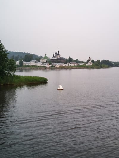 The image captures a serene view of the Goritsy Monastery, also known as the Resurrection Monastery, located on the shores of Lake Kubenskoye in Goritsy, Russia. The monastery complex, characterized by its white walls and numerous domed churches with green and black roofs, stands prominently against a hazy sky. A bell tower with a white facade and dark dome is visible on the right side of the complex. In the foreground, the calm, dark waters of the lake stretch towards the monastery. A white buoy with the number "79" printed in orange is positioned in the middle of the lake, serving as a navigational marker. The left side of the image features a lush green bank of reeds extending into the water, with trees forming a backdrop to the monastery. The overall atmosphere is tranquil and overcast, suggesting a diffused light. There are no people visible in the frame.