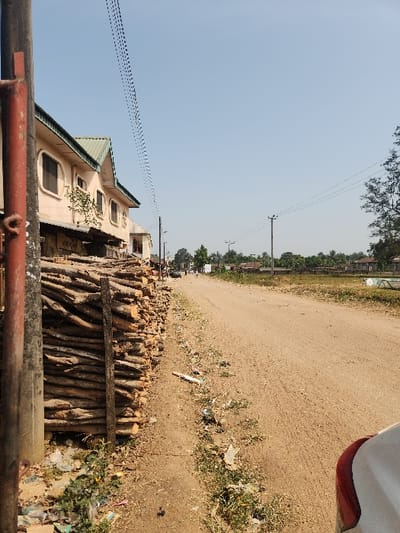 This image captures a bright, sunny outdoor scene, likely in a semi-rural or developing area. The main feature is a wide, unpaved dirt road or pathway extending into the distance, flanked by various elements.
On the left side of the road, a light-colored building, possibly cream or light yellow, with a dark green gabled roof stands prominently. It features several distinct windows with rounded tops. Overhead, a clear, pale blue sky dominates the upper portion of the frame, crisscrossed by numerous utility lines stretching between power poles that recede into the background. Distant trees and faint structures are visible along the horizon line.
Along the right side of the road, a large, organized stack of cut logs or firewood is piled. The ground immediately adjacent to the road and the log pile is earthy, interspersed with patches of green scrub and scattered litter or debris. A small section of a white vehicle, displaying a red element (possibly a bumper or tail light), is visible on the far right edge of the frame. In the immediate foreground at the bottom, a dark metal fence or railing partially obstructs the view.
The scene is devoid of people, suggesting a quiet moment. The bright, clear sky and strong illumination indicate it is daytime, likely mid-morning or early afternoon, with sunny weather. No specific activity or event is occurring. There is no visible text in the image. The overall setting is consistent with a location such as Umuoye, Nigeria, featuring unpaved roads and common local architecture and resources like firewood.