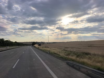 The image depicts a view from a highway in Wilsdruff, Germany, looking towards a rural landscape under a cloudy sky with the sun breaking through. The highway, made of asphalt, has multiple lanes marked with white lines. On the left side of the highway, there is a dense line of trees. To the right of the highway, a field of dry grass and crops stretches into the distance. Several wind turbines are visible on the horizon, scattered across the field. A metal guardrail is present along the right edge of the highway. The sky is filled with dramatic clouds, with rays of sunlight shining through in the upper right portion of the image. There are a few cars visible on the highway in the distance.