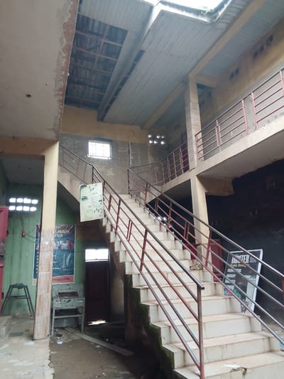 The image depicts the interior of a multi-story building with an open-air courtyard area. A prominent concrete staircase with metal railings ascends from the ground floor to the upper levels. The building appears to be under construction or in a state of disrepair, evidenced by the exposed ceiling beams and corrugated metal roofing in some areas.
On the ground floor, posters are displayed on the walls. One poster advertises "AL HEALTH CENTER" with images of animals, likely related to veterinary services or agricultural products. Another poster, positioned on the staircase, features a woman's face and text related to cosmetics, with small circular images that could be product samples or ingredients. Further up the stairs, a poster for "UMTER" graphics and printing services is visible.
To the left of the staircase, a metal cutting machine is positioned next to a green wall with more posters. A simple metal stand is also present. The ground is covered with debris, including pieces of wood, plastic, and what appears to be a used tire. Water has accumulated in some areas, suggesting a lack of proper drainage or recent rainfall. The overall environment is one of a semi-urban setting, possibly a commercial or community building in Mbar, Nigeria, that is either in its early stages of development or has been neglected.