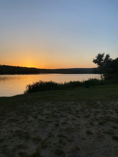 This is a horizontal, eye-level, full shot image of a sunset over a lake in Găleștii Noi, Moldova. The sky is a gradient of pale blue at the top, transitioning to a bright orange near the horizon. The sun is low on the horizon, partially obscured by a distant, dark, tree-lined hill. The lake fills the middle ground of the image, reflecting the warm hues of the sunset. The water is calm with small ripples. In the foreground, there is a grassy embankment that slopes down towards the water. Tall reeds and grass grow along the edge of the lake, silhouetted against the water. A single dark green trash can is visible on the grass to the right of the reeds. The overall mood of the image is serene and peaceful.