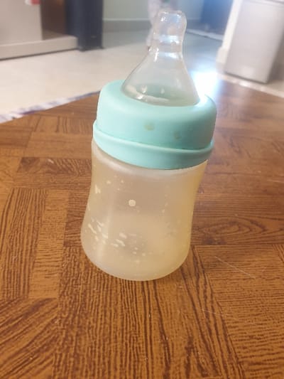 A translucent, frosted plastic baby bottle with a light teal cap and clear silicone nipple stands upright on a brown, wood-patterned floor. The bottle contains a residual amount of light yellow liquid with white particulate matter. Small liquid droplets are visible on the exterior of the teal cap. The background is out of focus, revealing a blurred indoor environment with light-colored flooring and indistinct furniture or appliances. This scene is located in Mumbuni North ward, Kenya.