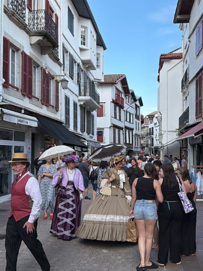 An outdoor scene captures a crowd of people gathered on a cobblestone street, lined with buildings, likely in the city of Saint-Jean-de-Luz, France. The buildings have white facades and are accented with red and dark-colored shutters. Some individuals are dressed in period costumes, including a man in a striped suit and straw hat, and women in long dresses, one holding a white lace parasol. Mixed among them are modernly dressed tourists. The sky is visible above the buildings, and the angle of the shot suggests the photo was taken with a mobile phone or similar device, possibly held at an unusual angle.