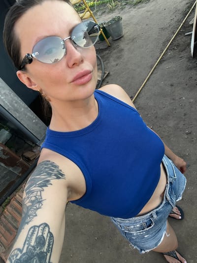 A woman with dark hair pulled back and wearing sunglasses, a blue tank top, and denim shorts is standing outside on a dirt path. She has tattoos on her left arm. In the background, there is a metal fence, some bricks, and plants.