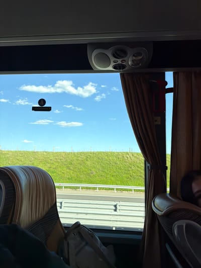 The image appears to be taken from inside a moving vehicle, potentially a bus or coach. The window view includes a blue sky with scattered clouds, a green grassy area, and a highway with a guardrail. Inside the vehicle, there is a speaker system mounted on a panel, brown curtains partially drawn, and tan upholstered seats. A sign written in German, possibly indicating an emergency exit, is visible. This does not provide enough information to place this photo in Malko Tranovo, Bulgaria.
