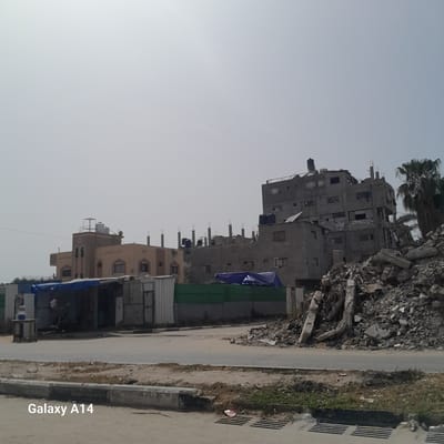 The image shows a street scene in the city of Nuseirat, Palestinian Territories. A partially destroyed gray multi-story building is visible, with a pile of rubble next to it. Other buildings, some appearing intact, are also present. The street itself appears paved, with a curb and some vegetation along the side. There is also what seems to be a storm drain visible. The sky is overcast and the lighting is soft. The text "Galaxy A14" appears in the bottom right corner.