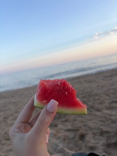 A close-up shot features a slice of watermelon being held by a person's hand. The watermelon is bright red with some seeds visible, and a sliver of green rind. The person's hand has light pink polished nails and a delicate silver ring on their finger. In the background, the scene transitions from a sandy beach to the ocean, with a soft, diffused light suggesting it might be during the golden hour, either sunrise or sunset. The sky is a pale blue, blending into soft, warm tones at the horizon. The overall mood is relaxed and serene, possibly indicating a leisurely moment at the beach. The sandy texture of the beach is visible in the upper right portion of the image, with some darker patches and slight elevation changes, perhaps suggesting footprints or natural undulations. The water appears calm, with a gentle transition from wet sand to the sea. The setting is consistent with a beach location, and the presence of the watermelon suggests a refreshing snack enjoyed outdoors.