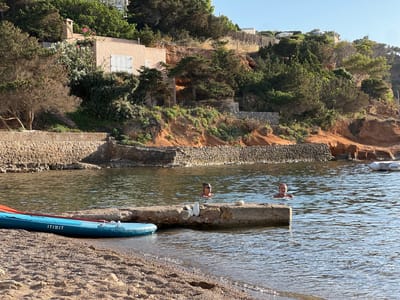 The image depicts a serene coastal scene in Sant Josep de sa Talaia, Spain. The foreground features a pebbly beach with a blue kayak resting on its side, partially submerged in clear, calm water. The kayak has an orange paddle attached and the brand name "ITIWIT" is visible.

In the middle ground, two people are swimming in the water. One person, wearing sunglasses, is partially submerged and facing the viewer. Another person, also in the water, is facing left. They are near a concrete jetty that extends into the water. A pair of white sandals are placed on the jetty.

The background reveals a lush green hillside dotted with trees and shrubs. A light-colored building with white shutters is nestled into the hillside, suggesting a residential or holiday property. Stone walls line the shoreline. The overall impression is one of a peaceful, sunny day, likely in the late afternoon or early morning given the quality of the light. The water is calm and reflects the blue sky. The natural landscape is enhanced by the presence of human activity, creating a harmonious blend of nature and recreation.