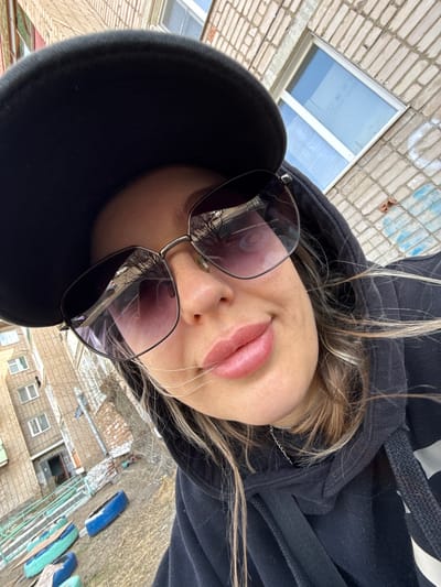 A downward-angled selfie of a person wearing sunglasses, a black hoodie, and a black baseball cap. The individual has blonde hair and full lips. The sunglasses reflect a scene with brick buildings. The background includes a brick building facade with a window, and another building structure is visible further down. A blue object (possibly a car) is discernible in the lower right corner, next to something made with wooden planks. This seems to be a view from a height, possibly a balcony or roof. Without further information, I can't confirm the exact location in Borzya, Russia.