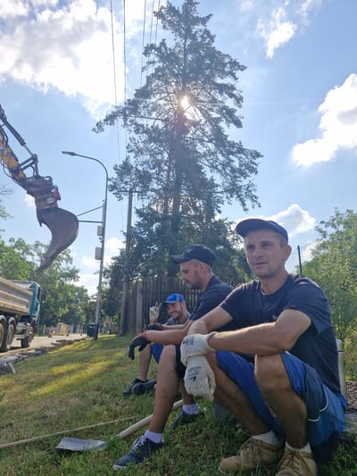 In Střelice, Czechia, on a sunny day, three men are taking a break from their work on a construction site. An excavator's bucket is visible in the upper left corner, and a dump truck is parked nearby. Paving stones are stacked on the side of the road, suggesting that the work involves road construction or repair. The men are dressed in casual work clothes, and two of them are wearing baseball caps. They are sitting on the grass, which is slightly overgrown in some areas. A tall pine tree dominates the background, with its branches reaching towards the bright blue sky filled with scattered clouds. Power lines stretch across the frame, and a street lamp stands tall beside the road. The overall scene suggests a moment of rest during manual labor in an outdoor, semi-urban environment.