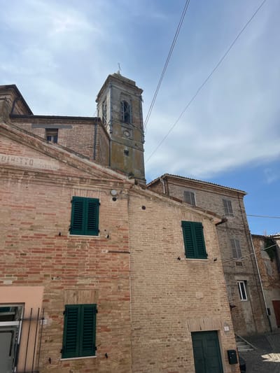 The image depicts a view of buildings in the town of Barbara, Italy, with a bell tower visible in the background. The foreground features a brick building with dark green shutters on its windows. To the right, another brick building with similar shutters is visible, along with a doorway and a set of stairs. The sky above is blue with scattered clouds. The scene appears to be an exterior shot of a street or alleyway within the town.