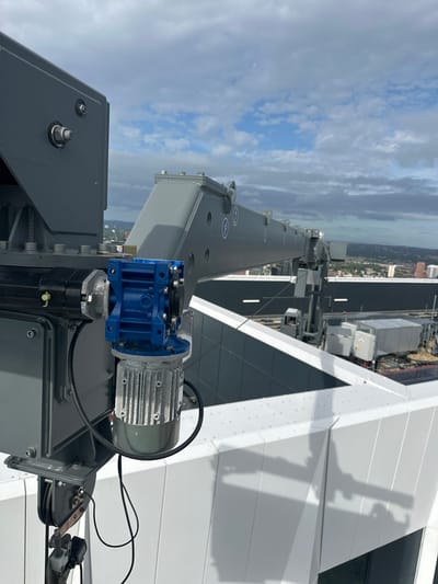The image shows a close-up view of a crane mechanism, likely on a rooftop in Birmingham, United Kingdom. The crane arm is a light grey, industrial metal structure extending into the distance. Attached to the main support structure is a bright blue gearbox and a silver, ribbed electric motor. The motor is connected to the gearbox by a shaft. Below this assembly, a pulley system with a steel cable is visible, suggesting the crane is functional. In the background, more of the crane's structure can be seen, along with other rooftop equipment and the cityscape of Birmingham under a partly cloudy sky. The foreground features a white, modern architectural element, possibly part of the building's facade or rooftop.