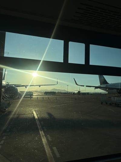 An airport tarmac is seen through a window with distinct panes. Several aircraft are visible, some parked and others possibly taxiing. The sun is bright, creating a strong lens flare and casting shadows on the ground. Mountains are faintly visible in the distance. The ground appears to be concrete or asphalt, marked with white lines.
