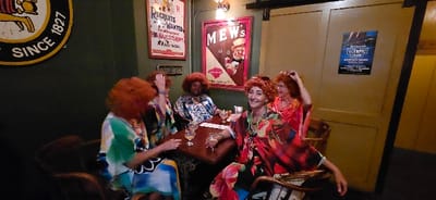 The image displays five adult females seated around a dark wooden square table inside an establishment, identified by a poster as The Centralia Olympic Club in Centralia, Pennsylvania, United States. All five subjects are wearing identical vibrant orange, curly wigs and distinctive, brightly colored, patterned, loose-fitting tops or kaftans. The female in the foreground, seated center-right, is smiling directly at the camera while holding a clear glass containing a light-colored beverage. Another female across the table from her also holds a similar glass. Two additional beverages and a small white napkin are present on the table surface. The background features dark green walls to the left, adorned with three framed items: a large circular yellow and black sign bearing "SINCE 1827", a red-framed poster titled "RECRUITS WANTED FOR HER MAJESTY'S ARMY", and a red-framed advertisement for "MEW's". To the right, a lighter wooden wall displays a black and blue poster advertising "The Centralia Olympic Club Monthly Picnic". Overhead lighting is provided by an exposed incandescent bulb fixture on the left wall.