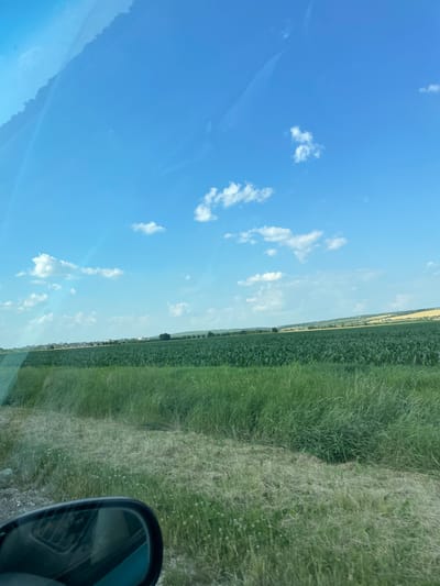 The image depicts a rural landscape under a bright blue sky with scattered white clouds. In the foreground, a grassy verge and a gravelly surface are visible, with a side-view mirror of a vehicle partially in the bottom left corner. Behind the verge, a vast field of green crops, likely corn, stretches across the mid-ground. In the distance, a line of trees and buildings forming a settlement can be seen, suggesting the outskirts of a town or village. The horizon is marked by rolling hills, some of which appear to be cultivated with yellow crops. The overall scene is serene and agricultural. The metadata indicates the location is Valea Mare, Moldova. There are no people or animals visible in the image, and no apparent actions or interactions taking place.