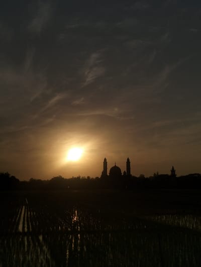 A silhouetted mosque stands against a bright, sunlit sky with dark clouds above. The sun casts a golden glow around the structure. In the foreground, a field of dark vegetation, possibly rice paddies, reflects some of the light. The overall mood is tranquil and serene.