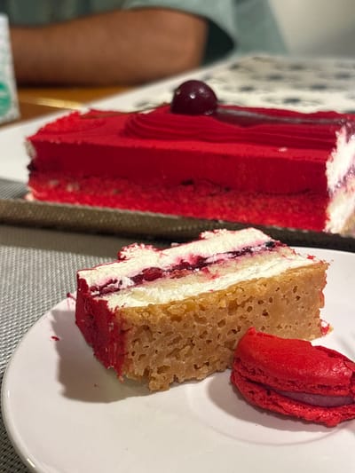 The image displays an indoor scene focused on desserts, captured at close range. In the foreground, a slice of a multi-layered cake and a single red macaron are presented on a white circular plate. The cake slice features a light brown cake base, topped by a thick white cream layer, followed by a thinner dark red berry or fruit compote layer, and a final white cream layer, with one edge showing a red crumb or velvet spray finish. Adjacent to the cake slice, the red macaron has a red shell and a discernible purple or dark pink cream filling. In the blurred mid-ground, a larger, partially consumed rectangular red cake is visible, from which the foreground slice appears to have been cut. This cake is uniformly covered in smooth red frosting with a decorative piped red border and is garnished with a single dark red cherry or berry on top. Both the main cake and the plate are situated on a light grey textured placemat or table covering. In the soft-focus background, a person's arm or upper torso, clad in a light blue-green shirt, is partially visible, along with a small, blurred white object featuring green text, possibly a package or container. The specific location within Bnei Dror, Israel, cannot be visually confirmed from the details present in the image.