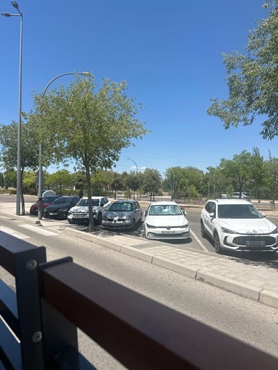 The image shows a row of parked cars on a sunny day in Húmera-somosaguas-prado Del Rey, Spain. The cars are parked parallel to a sidewalk and a street. There are several trees casting shadows on the ground. The sky is clear and blue. The cars visible include a red Seat, a black Seat, a white Volkswagen Golf, and a white MG. The license plates are visible on some of the cars. In the foreground, a dark railing is partially visible, casting a shadow across the street and sidewalk. The environment appears to be a suburban or commercial area with infrastructure like streetlights and power lines in the background. No people are visible in the image.