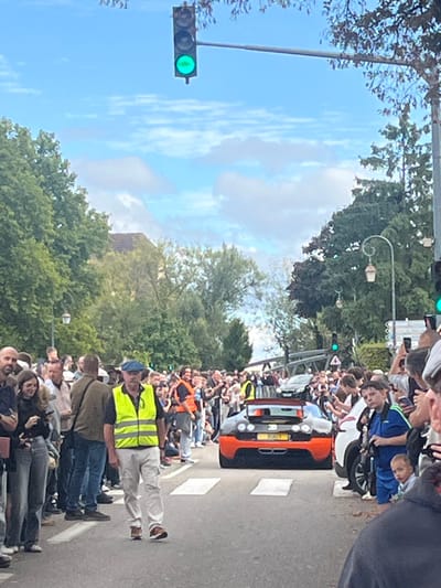 An outdoor scene in Molsheim, France, depicts a black and orange Bugatti Veyron-like vehicle moving away from the viewer on a paved road. The vehicle, featuring a prominent rear wing and a partially visible license plate displaying "BJY", is positioned near the center of the street. White crosswalk stripes are visible on the asphalt in the foreground.

Dense crowds of spectators line both sides of the road, extending into the background, observing the vehicle. Many individuals within the crowd are holding up smartphones, indicating photography or video recording.

In the immediate foreground, slightly to the left of the road's center, a man walks towards the viewer. He wears a blue flat cap, a neon yellow high-visibility vest with reflective stripes, a light-colored shirt, light-colored pants, and brown shoes. He possesses a mustache.

The environment includes green trees lining both sides of the street behind the crowds. A building with a reddish-brown roof is partially visible through the trees on the left mid-ground. Decorative streetlights with curved arms and lantern-style fixtures are present further down the street. Overhead, two traffic lights display green. The sky is partly cloudy with visible blue areas. A bridge-like structure is discernible spanning the street in the background.