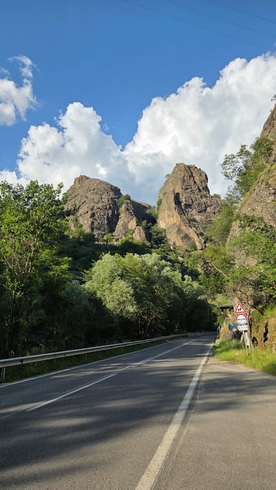 The image depicts a scenic mountain road in the vicinity of Rila, Bulgaria. The road, paved and marked with white lines, curves gently to the right, bordered by a metal guardrail on the left and lush green foliage on the right. Towering rock formations, weathered and rugged, dominate the background, with trees and shrubs clinging to their slopes. The sky is a vibrant blue, adorned with fluffy white clouds. Several road signs are visible on the right side of the road, indicating a sharp curve ahead and potentially other traffic regulations. The overall impression is one of a tranquil, natural landscape, likely a popular route for scenic drives or outdoor excursions in the Rila region.
