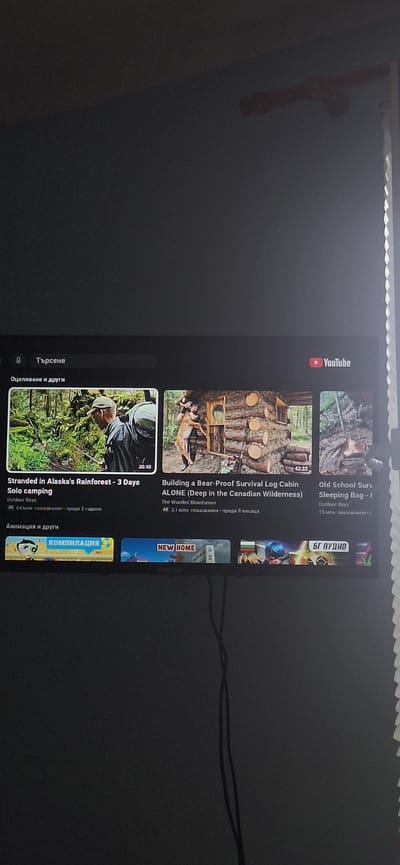The image displays a television screen showing the YouTube interface. The interface is displaying a grid of video thumbnails under the category "Оцеляване и други" (Survival and others). The top left corner shows a search bar with the text "Търсене" (Search).
One prominent video thumbnail shows a person with a backpack in a lush green forest, with the title "Stranded In Alaska's Rainforest - 3 Days" and "Solo camping" below. Another thumbnail features a person holding a deer in front of a log cabin, titled "Building a Bear-Proof Survival Log Cabin ALONE (Deep in the Canadian Wilderness)". A third thumbnail shows a person by a campfire, with the title "Old School Surv... Sleeping Bag - I". Below these, there are additional thumbnails with titles like "КОМПИЛАЦИЯ" (Compilation) and "NEW HOME". The YouTube logo is visible in the top right corner.
The television is mounted on a dark wall. A white, textured object, possibly a decorative element or part of the television frame, is visible on the far right edge of the image. Two black cables are hanging down the wall in the lower right quadrant of the image.
There is no information in the image that directly places the scene in Ovcha Mogila, Bulgaria. The text visible is in Bulgarian.