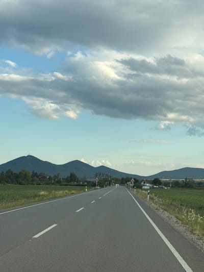 The image shows a two-lane asphalt road leading towards a mountain range under a partly cloudy sky. The road has white lane markings. On the left side of the road, there are green fields and trees. On the right side, there are also green fields, some with tall grass and wildflowers, and a few houses or buildings in the distance. Several traffic signs are visible along the right side of the road, including warning signs. The mountains in the background are dark green and some have a hazy appearance. The sky is a mix of blue and grey clouds, with sunlight breaking through in some areas. The scene is one of a rural landscape in Luhyňa, Slovakia.