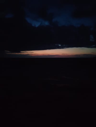 A wide shot captures the dark silhouette of the Russian countryside at twilight. The sky is a deep indigo, with ominous, heavy clouds gathering. Near the horizon, a faint band of pale orange and pink light indicates the last remnants of daylight, with a few smaller clouds scattered within it. The foreground is entirely black, suggesting a landscape devoid of any light sources or visible features. The composition is dominated by the dark expanse of the land and the dramatic, cloud-filled sky.