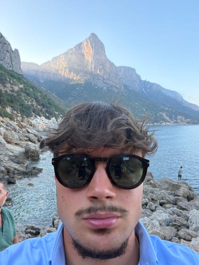 This is a selfie taken by a young man with dark, tousled hair and a mustache and goatee. He is wearing dark sunglasses and a light blue collared shirt. He is in the foreground, with a body of water and a rocky coastline behind him. In the middle ground, the water stretches out to the horizon, with a person standing on the rocks on the right. In the background, there are steep, verdant mountains. The scene is lit by what appears to be late afternoon sun. The location is the coast of Baunei, Sardinia, Italy, a municipality known for its rugged coastline and dramatic mountain scenery.