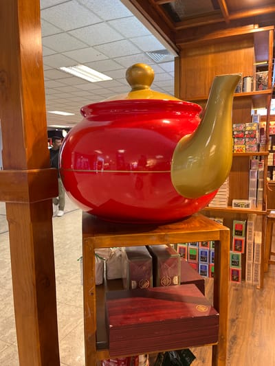 An oversized, glossy red teapot with a golden lid and spout is displayed on a wooden shelf in what appears to be a retail setting. The teapot is positioned prominently in the foreground, dominating the frame. Behind it, shelves are stocked with various boxes, likely containing tea, given the context of the teapot. The shelves are made of polished wood, and the overall aesthetic suggests a shop that specializes in tea or related products. In the background, a ceiling with fluorescent lighting and ceiling tiles is visible, indicative of an indoor commercial space. A man in casual attire, a dark t-shirt and light-colored trousers, is walking in the background, slightly out of focus, suggesting this location might be a commercial area or airport in Katunayake, Sri Lanka, possibly a duty-free shop or a specialty store. The lighting is bright, illuminating the products and the environment.